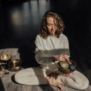 Female sound therapist sitting with crossed legs on parquet while playing singing bowl with mallet during meditation practice